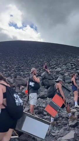 The video begins with a group of young adults on a rocky, dark-colored slope, likely volcanic ash or scree. The sky is overcast with dramatic, grey clouds, though hints of blue sky are visible. The overall impression is of a challenging hike under a moody sky. The people are dressed casually, ready for outdoor activity, with backpacks and water bottles. One woman in the foreground wears a black backpack with white text that reads "MEDIUM" and below that "LOS LAURELES".

The camera pans across the landscape, revealing a vast, steep, dark slope that ascends into the cloudy sky. The texture of the ground is rough and uneven, composed of countless small, dark rocks. The terrain suggests a volcanic environment.

As the video progresses, the sky begins to clear, revealing a brighter, more expansive blue sky with fluffy white clouds. The camera pulls back to reveal more of the setting. A lush, green hillside slopes down into the distance, contrasting sharply with the dark volcanic material. The scenery suggests a transition from a barren volcanic area to a more vegetated landscape. The sunlight breaks through the clouds, casting dappled light on the green hills. The final shots offer a wide panorama of rolling green hills under a bright, partly cloudy sky, with the dark, rocky foreground still prominent. The overall mood shifts from a somewhat somber, challenging hike to one of scenic beauty and awe as the weather clears and the landscape opens up. The location is likely a volcanic area in Rota, Nicaragua, given the distinct contrast between volcanic rock and lush greenery.