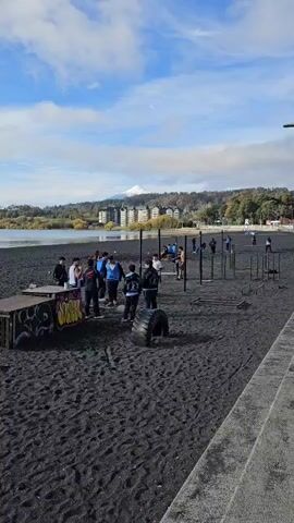 The video shows a group of people exercising on a black sand beach on a partly cloudy day. There are about 15 people, mostly young men, wearing athletic clothes and some wearing jackets. Some are gathered near a wooden platform with graffiti, while others are using pull-up bars and other exercise equipment. A large tire is also present. In the background, there is a calm body of water with buildings lining the shore, and a snow-capped mountain is visible in the distance. The scene appears to be a beach workout session. The camera slowly zooms in on the group of young men.