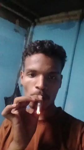 The video is a first-person perspective of a young man smoking a cigarette. The man is holding a lit cigarette between his fingers and exhaling smoke. He is wearing a brown t-shirt and has curly dark hair. The background appears to be an indoor setting with a blue wall and some dark objects. The video is short and appears to be a casual recording.