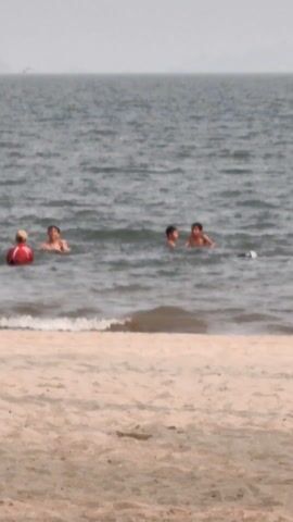 The video shows a beach scene. The foreground is a sandy beach, and beyond that, the water stretches out to the horizon. In the water, there are several people swimming and interacting. Some appear to be kids, while others seem to be adults. They are spread out in the water, some closer to the shore and others further out. The water is a dark grey, and the sky is overcast, creating a slightly hazy look. In the distance, there are some land masses or islands visible.