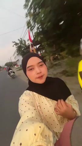 A woman wearing a black hijab and a cream-colored dress with brown polka dots is filmed from a first-person perspective, possibly from a motorcycle or scooter. She is looking at the camera and making a kissing gesture. In the background, there is a road with trees and a few other motorcycles. The lighting suggests it is either early morning or late evening. The video is shaky, indicating movement.