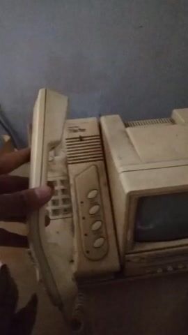The video shows a hand picking up a phone handset from a dusty, beige intercom system. The system includes a phone base with buttons, and a small television monitor. The backdrop is a plain blue wall. The hand holds the handset and briefly says, "Hello."
