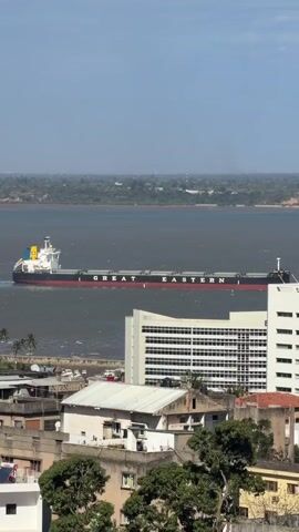 A large cargo ship named "GREAT EASTERN" sails across a wide body of water. The ship is long and black with a white superstructure and a red stripe along its hull. It has a yellow and blue funnel. In the foreground, there are several buildings, including a tall, modern white building with many windows, and other smaller, older buildings with tiled roofs. Trees are visible in front of some of the buildings. The background shows a distant green coastline under a clear blue sky. The water appears choppy, indicating some movement or wind. The perspective suggests the shot is taken from an elevated position, looking down and across the scene.