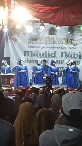 A group of women in blue dresses and headscarves are singing on a stage in front of a large banner. The banner says "PENGAJIAN UMUM Dalam Rangka Tasyakuran & Peringatan Maulid Nabi Muhammad SAW 1440 H". There is a crowd of people watching them from below. The stage is lit with bright lights.