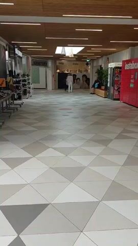 A panning shot moves across a retail environment, possibly a mall or shopping center. The floor is tiled in a geometric pattern of light grey, dark grey, and off-white triangles. Various shops and displays are visible along the sides. On the left, a shoe store is apparent with shelves filled with footwear. In the foreground, tables with shoes are displayed. In the background, people are seen walking and browsing. One shop sign reads "SKOKJÆR". Another sign indicates "Center Kiosken". A popcorn cart is also visible. The overall impression is a well-lit, modern indoor shopping area.