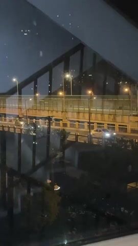A nighttime scene captures a bridge illuminated by streetlights. Cars travel across the bridge, their headlights and taillights visible as streaks of light. The reflection of the bridge and the sky can be seen in a glass surface in the foreground, which also shows a dark, angled structure that might be part of the building the camera is in. Trees are visible below the bridge, their shapes silhouetted against the night. The overall impression is of an urban environment at night, with the constant movement of traffic.