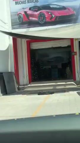 The video shows a street scene in front of a building, with signage in Spanish including the name "Dr. Simi". The building has a red and white color scheme, and there are several parked cars visible. A person wearing a white costume is walking on the sidewalk in front of the building.
