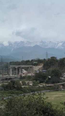 The video presents a landscape with a bridge under construction in the foreground, set against a backdrop of snow-capped mountains. The sky is overcast, casting a muted light over the scene. The bridge appears to be in the early stages of construction, with concrete pillars and some segments of the deck already in place. Construction vehicles and equipment are visible around the site. In the middle ground, there is a green field or park. Trees and vegetation fill the spaces between the construction site and the field, partially obscuring the view. Power lines and utility poles are visible, adding to the infrastructure elements in the landscape. The camera angle is from an elevated position, providing a comprehensive view of the scene.