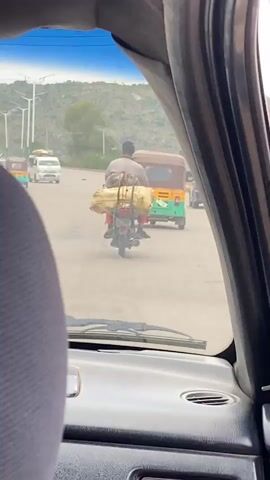 The video captures a view from inside a vehicle, looking out through the front windshield and side window. The main focus is a person riding a motorcycle loaded with large, yellow-wrapped bundles secured to the back. The motorcycle is moving forward on a paved road. Behind the motorcycle, an auto-rickshaw, also carrying a passenger or goods, is visible. In the background, there are trees, hills, and utility poles under a bright sky. The overall scene appears to be a street or road in a somewhat rural or developing area, with traffic and everyday activities taking place.