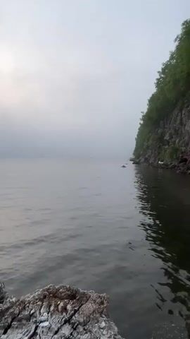 A vertical video shows a calm lake under a hazy sky. In the foreground, a rough rock is visible on the left. The water extends to the horizon, where it meets the hazy sky. On the right side of the frame, a steep, rocky cliff covered in green trees rises from the water's edge. The water is dark and reflects the sky and the cliff. There are a few small objects floating on the water, and a white object, possibly a bird or a sign, is visible on the rocky shore near the cliff. The scene appears to be an outdoor, natural environment.