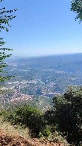 This is a video taken from a high vantage point, looking down on a valley. The valley contains a river, a town or city, roads, bridges, and buildings. The terrain is mountainous and covered in trees and vegetation. The sky is clear and blue. In the foreground, there are trees and some dirt or rocky ground. The camera pans slightly, showing different parts of the valley.