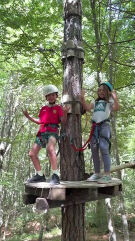 Two children, a boy and a girl, stand on separate wooden platforms attached to a tree trunk in a lush green forest. Both children wear safety harnesses and helmets. The boy is on the left, wearing a red shirt, green shorts, and a white helmet. He poses with both hands forming a "shaka" sign. The girl is on the right, wearing a white t-shirt, grey pants, and a light blue helmet. She also forms a "shaka" sign with both hands. They both appear to be happy and smiling. The video pans down, revealing more of the forest floor and other rope courses in the background.
