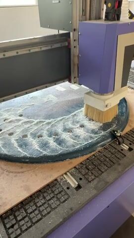 The video shows a CNC machine cutting into a piece of blue plastic. The machine, which is primarily purple and silver, has a white brush attached to its head. The brush rotates and disperses white plastic shavings as the machine cuts the blue plastic. The blue plastic is held in place by metal clamps on a wooden surface. There are several holes already cut into the blue plastic. The machine moves slowly and methodically. The environment is a workshop or factory setting.