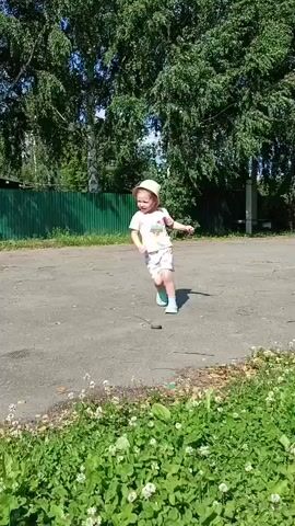 A young child wearing a pink t-shirt, white shorts with a pattern, a light-colored hat, and turquoise shoes stands on an asphalt path. The child is outdoors, with a green fence and trees visible in the background. The child is in motion, running and then stopping. The child appears to be speaking or yelling.