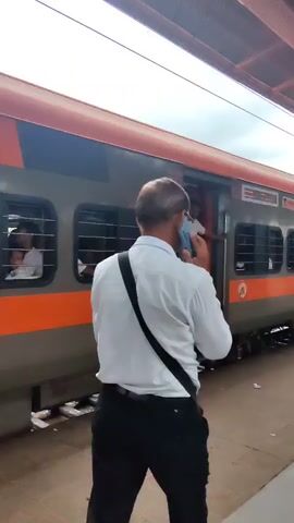 The video shows the side of a train at a station. The train car has gray and orange coloring. There is an open doorway with a few people standing near it. One person in a white shirt and dark pants is standing in the doorway, looking towards the camera. Another person in a red shirt and blue jeans is also in the doorway, with their arm raised. Two other people are visible through the windows of the train car. One is in the foreground, wearing a white shirt and holding a dark object, likely a phone or wallet, in their right hand. The train car has "SECOND CLASS" written on it in large letters. The environment appears to be a train platform, with tracks visible below the train.