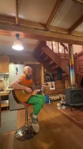 A man is sitting on a red stool playing a classical guitar in a rustic log cabin. The man has a beard and is wearing an orange tank top and green and black striped pants with furry slippers. The cabin has a wooden floor, a wood stove, and a staircase leading to a loft. There are fairy lights strung around the cabin. A kitchen area with a refrigerator is partially visible to the left of the man. The man is singing while playing the guitar.