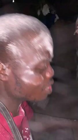 A close-up shot shows a man’s head and shoulders. He is dark-skinned, with a short beard. The top of his head is covered in a white powder. He wears a red shirt, and a strap hangs across his shoulder. He is looking to the side and appears to be speaking. The background is dark and indistinct, with glimpses of other people and objects.