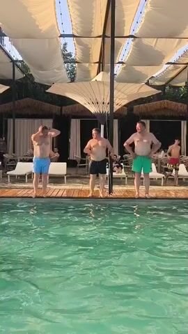 Three men are standing on a wooden deck next to a swimming pool. The man on the left is wearing blue swim trunks, the man in the middle is wearing black swim trunks, and the man on the right is wearing green swim trunks. The man on the left has his hands on his head. The man in the middle has his hands on his stomach. The man on the right has his hands on his hips. The swimming pool is filled with clear blue water. Behind the men, there are lounge chairs and umbrellas. The scene appears to be a resort or a private backyard.