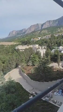 The video begins with a view of a large building complex surrounded by trees, a winding road, and mountains in the background. The camera then pans upwards, passing a scaffolding pole, to reveal a broader landscape. This includes the tree-covered terrain sloping towards the sea. We see the sea stretching out to the horizon under a partly cloudy sky. At one point, two people can be seen walking near the treeline. The footage appears to be captured from an elevated position, potentially from a high building or structure under construction.  There also seems to be construction equipment operating near the winding road.