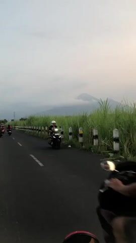 The video shows a side view of several motorcycles riding on a paved road. The camera is positioned behind the lead motorcycle, which has two people on it. The driver is wearing a dark shirt and black pants, and the passenger, wearing a teal headscarf and a green shirt, is seated behind them. To the right, a second motorcycle follows closely, with a person wearing a black helmet and white shirt. In the distance, a mountain peak is visible above a field of tall grass. The sky is overcast.