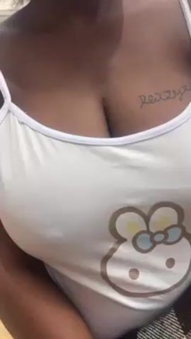 A close-up shot shows the upper torso of a person with dark skin, wearing a white tank top with a light brown cartoon bear-like face printed on it. The tank top has white straps and trim. A tattoo of a word is visible on the person’s right collarbone area. The background is out of focus, but appears to be outdoors with hints of green vegetation. The person remains still throughout the video. The lighting appears to be natural daylight.