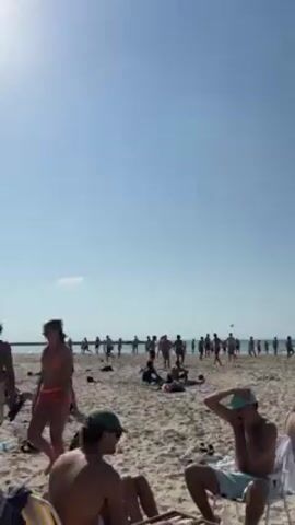 The video shows a sunny beach scene with a large group of young people playing a game involving multiple volleyballs. Many of the players appear to be men in swimwear, actively throwing and hitting the balls. Some people are sitting or lying on the sand, some in chairs, watching the game. The sky is clear and blue, and the sand appears light in color. A breakwater is visible in the background along the horizon.
