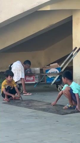 Three barefoot children are playing on a concrete slab under a partially visible building or structure. One child is squatting, holding a metal tool near the slab. Another child is squatting nearby, reaching with their hand towards the slab. A third child is standing behind a hand truck filled with cardboard boxes, looking into one of the boxes. The hand truck is positioned behind and between the two children on the slab. The lighting suggests it is daytime, and the overall environment appears simple and slightly rundown.