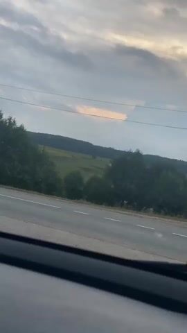 The video shows a view from inside a vehicle looking out at a rural landscape. The camera is positioned behind the windshield, with the dashboard visible in the foreground. Outside, there is a road with a white line down the center, bordered by trees and fields. Several tall, triangular power poles with electrical wires stretch across the frame. The sky is cloudy, with a hint of orange light suggesting either sunrise or sunset. The vehicle appears to be in motion, as the landscape shifts slightly throughout the clip.