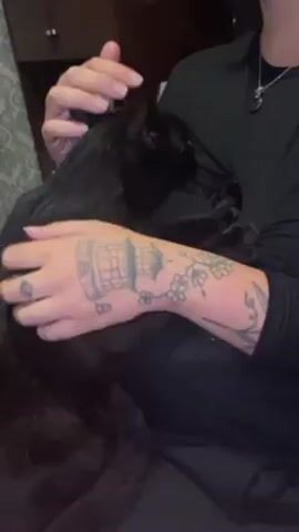 A person, wearing a long-sleeved black shirt and a pendant necklace, sits and pets a black cat. The person’s left arm is heavily tattooed. The background is a dark room with a cabinet visible in the background. The person pets the cat’s head, scratching the back of its ears.