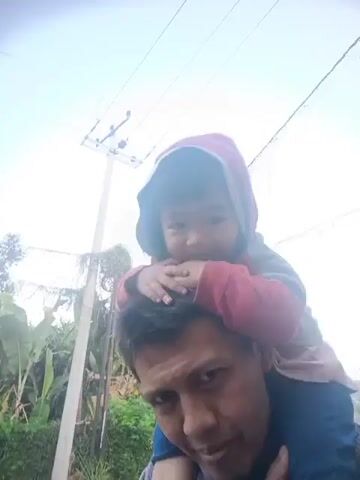 A video shows a person carrying a child on their shoulders while walking outdoors. The person is facing the camera, while the child is looking away. There are trees and a utility pole in the background. The video is grainy and appears to be taken with a mobile phone.