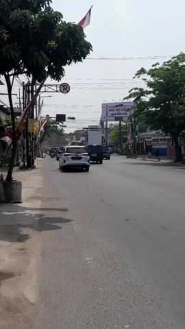 The video shows a street scene with several vehicles, including cars and motorcycles, driving on a road. There are buildings and trees lining the street, and a billboard is visible in the distance. The sky is clear and the weather appears to be sunny. The video is filmed from a stationary position on the side of the road, looking down the street.
