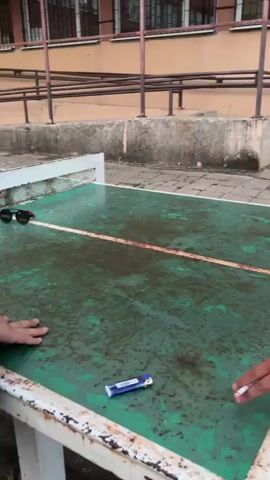 A short video shows a person attempting to perform a water bottle flip challenge. The person is standing to the right of a green and white outdoor ping pong table. They are wearing a black shirt and a dark vest. On the table, there is a pair of sunglasses and a blue cigarette lighter. The person holds a plastic water bottle and attempts to flip it so it lands upright on the table. The bottle lands on its side. The person then picks up the bottle and attempts the flip again, but the video cuts off before the result of the second attempt. The background shows a building with windows and a fence.