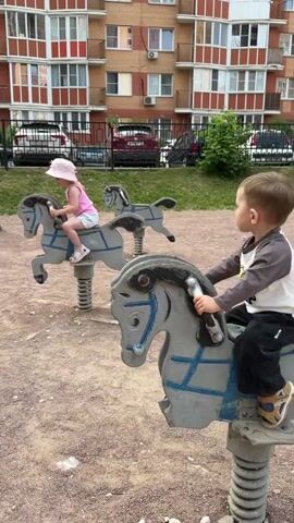 A mother is shown interacting with her young daughter who is riding a spring-loaded horse toy on a playground. The daughter, wearing a pink hat and shirt, is on one horse while another child, a boy, is on a similar horse in the foreground. The mother, with blonde hair, is reaching towards the daughter, possibly to help her. The playground has a sandy surface, and a brick apartment building is visible in the background, along with parked cars. The video appears to be captured outdoors during the daytime.