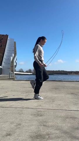 A woman with a ponytail is jump roping outdoors on a bright day. The area is a concrete walkway near a body of water, with a modern architectural building visible in the background to the left. She is wearing a light-colored zip-up jacket, black pants, white socks, and white shoes. The sky is blue and clear with a few wispy clouds. The water has some land visible across it. The jump rope appears to be a dark color. After a short jump roping session, she briefly stops.