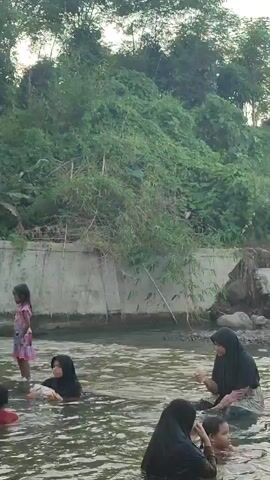 A group of people are in shallow water. In the foreground, a man wearing a striped t-shirt is partially visible, looking towards the camera. In the mid-ground, a woman wearing a black hijab is interacting with a child. To the left of them, another woman in a black hijab is partially submerged in the water. Further to the left, a young girl in a pink dress stands near the bank. In the background, a man in a black shirt is visible in the water. The environment appears to be a natural setting with lush greenery and a concrete structure along the water's edge. The water is murky and calm.