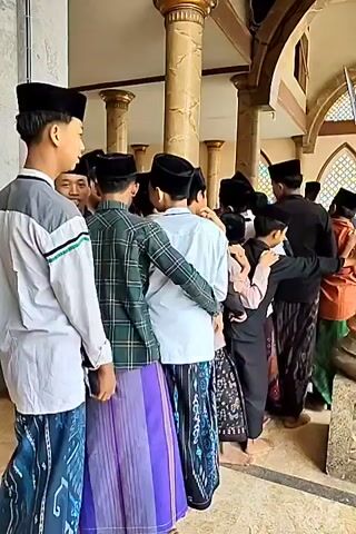 A group of young males, dressed in traditional Islamic attire including peci hats and sarongs, are standing in a line. They appear to be indoors, possibly in a mosque or a similar religious building, given the architectural elements such as large columns and patterned walls. The lighting suggests daytime. The young men are facing away from the camera, and some are interacting with each other, with one person's arm around another's shoulder. The camera pans slightly, showing more individuals in the line and the surrounding environment. The video then shifts to a wider view, showing the exterior of the building, with a street, cars, and other buildings visible in the background. The overall scene depicts a gathering of young men in a cultural or religious context.