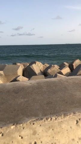 The video pans from a close-up of concrete tetrapods and a wall overlooking a calm ocean under a blue sky with scattered clouds. It then follows the concrete wall, showing a person sitting on it in the distance. The video continues to pan, revealing a sandy area leading towards modern buildings under construction, with the sun shining brightly in the sky.