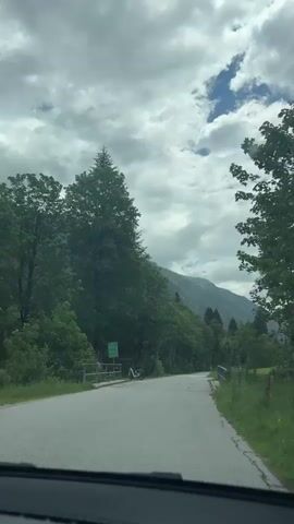 This is a first-person perspective video, likely filmed from inside a car, as it travels along a paved road. The scene is set in a picturesque mountainous region, with lush green trees lining both sides of the road. In the distance, majestic mountains rise, their peaks partially obscured by a cloudy sky. The weather appears to be overcast with some patches of blue sky and sunlight breaking through.

Along the left side of the road, a green signpost is visible, bearing text that is difficult to decipher due to the distance and resolution. A white bicycle is parked near the signpost. On the right side of the road, a wooden fence separates the road from a vibrant green grassy field. A rustic wooden building with visible windows is nestled in the field in the distance. The road itself has some puddles, suggesting recent rain.

The overall mood is serene and tranquil, evoking a sense of natural beauty and peaceful travel. The time of day is likely daytime, given the brightness of the scene. The location context of Flachauwinkl, Austria, suggests a scenic drive through alpine landscapes. There are no people clearly visible in the video. The activity is simply driving through this scenic environment.