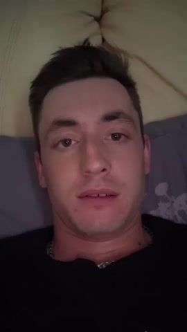 A man is lying down and speaking directly to the camera, presumably for a video. He is wearing a black shirt and a silver chain. The background appears to be a bed with a patterned pillow or blanket. He smiles at one point in the video.