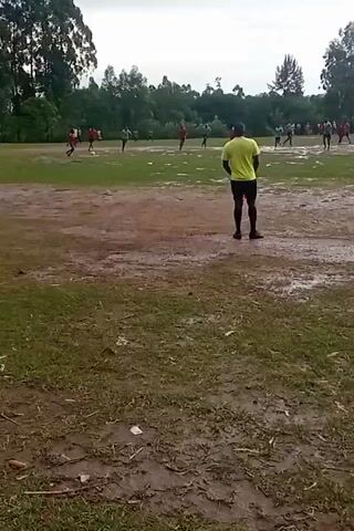 The video shows a soccer game in progress on a muddy field. A man in a yellow shirt and black shorts stands observing the game from a closer vantage point. The field is predominantly brown and muddy, interspersed with patches of green grass. The soccer players are scattered across the field, wearing red and white jerseys. The sky is overcast. Trees surround the field.