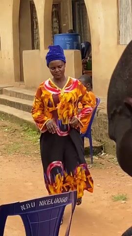 A woman in a colorful dress and blue headwrap stands in front of a building with steps. She is looking down and adjusting her clothing. To her left, a blue chair with writing on it is visible. Behind her, a man in an orange shirt sits on a blue chair. The environment appears to be outdoors with a dirt ground and a building made of what looks like mud or plaster. Another person's head is visible in the foreground, obscuring part of the scene.
