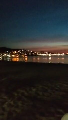 The video shows a beach at dusk or night. The sky is a deep blue with streaks of orange and red near the horizon, indicating the sun has recently set. In the distance, lights from a town or city glow along the coastline. The water is calm, reflecting the lights from the shore. The foreground is dark, showing a sandy beach and a white plastic chair. There are no people visible in the video.