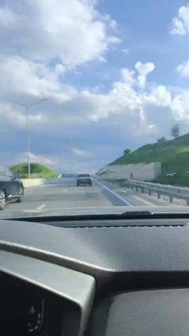 The video shows a first-person perspective from inside a moving car on a multi-lane highway. The dashboard of the car is visible at the bottom of the frame. In the distance, other cars are driving on the highway. To the left, there are streetlights and green hills. To the right, a retaining wall with green slopes above it is visible. The sky is partly cloudy with blue patches. A road sign on the right says "ПОДКУМОК" which is "PODKUMOK" in Cyrillic.
