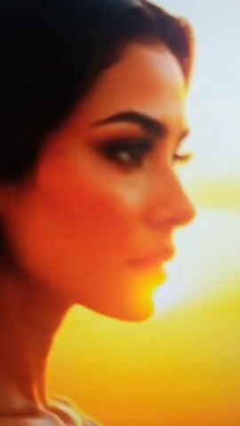 The video shows a woman with dark hair and striking makeup, looking to the right. She has a dramatic smokey eye and bold eyebrows. In the first few seconds, her face is overlaid with a semi-transparent, slightly distorted version of herself, also in profile. The background is a warm, glowing orange, suggestive of a sunset or golden light. As the video progresses, blue lightning-like graphics appear on her chest. Then, the scene shifts to a nighttime urban environment with a blurred cityscape and neon lights. A man in a dark suit and tie stands behind her, slightly out of focus. The woman is wearing a revealing black dress with intricate embellishments. Her hair is styled in loose waves.