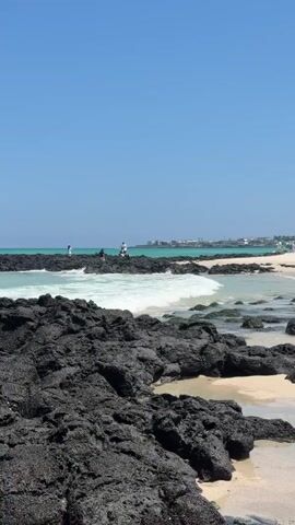 A vertical, eye-level shot of a beach scene on a sunny day. The foreground is dominated by dark, jagged volcanic rocks. Behind them, a gentle wave breaks on a sandy shore, revealing turquoise water. In the middle ground, several people are standing on similar volcanic rocks in the shallow water. One person, dressed in white, appears to be taking a photo. Further back, across the turquoise water, a town or resort with buildings is visible along the coast, with a clear blue sky above. The overall scene is serene and picturesque.