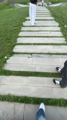 The video shows a person walking up a path of gray stepping stones interspersed with green grass. The path slopes slightly upward. The person is wearing blue jeans and white sneakers. Another person, wearing white pants and a brown jacket, is walking ahead on the same path. At the top of the path is a grassy area with a circular structure made of stone. Other people can be seen in the distance. The sky is partly cloudy. There are trees in the distance on the left. The camera is shaky and appears to be handheld. The person in the brown jacket turns to look at the camera and says something. The video abruptly cuts off after a few seconds.