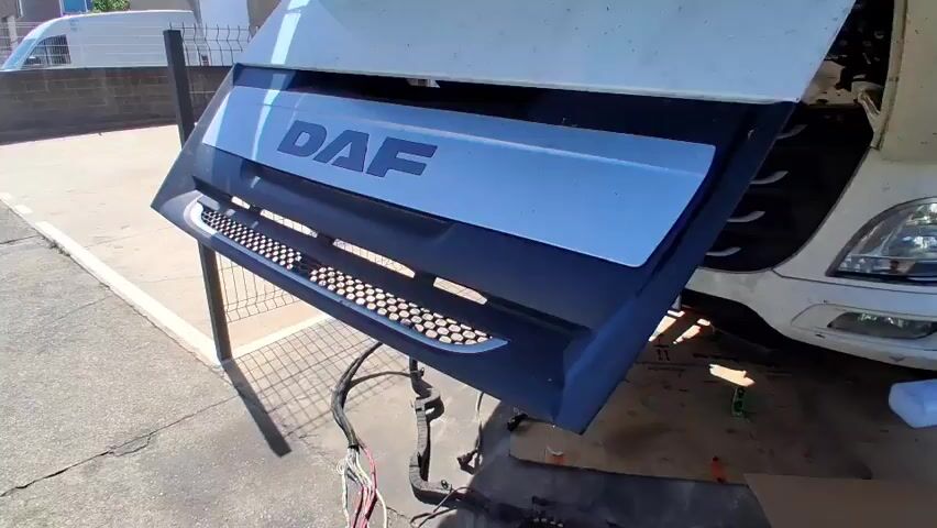 The video shows a close-up view of a DAF truck grille, which is propped up on a stand. The grille has the DAF logo clearly visible. In the background, a white truck is partially visible. The scene appears to be outdoors, possibly in a workshop or parking area, given the concrete ground and scattered cardboard boxes.

The primary activity seems to be the installation or removal of parts from the truck. A man in a black t-shirt with colorful squares and text that says "changing color values" is holding a complex wiring harness. Another person, wearing a black work outfit and a yellow safety harness, is seen in the background near a cardboard box. Wires and connectors are spread out on the ground, suggesting ongoing work.

The time of day appears to be daytime, with bright sunlight casting shadows. The weather seems to be clear. The license plate on the truck is visible and reads "CB 1529 KB." This indicates the vehicle is registered in Spain, and the mention of "Mas Aliu, Spain" in the prompt provides a specific location context. The overall mood appears to be functional, focusing on the mechanical task at hand.