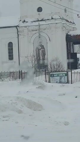 The video shows a white church building during a snowy day, viewed from inside a moving vehicle. The church has an image of a figure painted above an archway. A traffic sign with red and white chevrons is visible in front of the church, and there is a low metal fence separating the church from the road. The environment is snowy with a thick layer of snow covering the ground. The view from the vehicle includes parts of the car's interior, such as the windshield and side mirror.