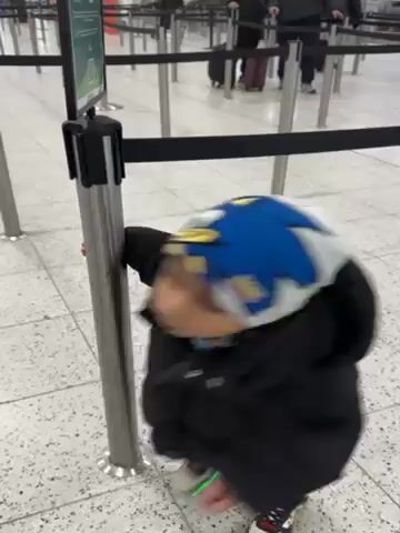 The video shows a young child, likely a toddler, playing near a metal stanchion at what appears to be an airport. The child is wearing a dark coat, blue jeans, and a patterned hat, and has light-up sneakers. The toddler interacts with the stanchion by walking around it, holding onto it, and then pushing away from it. The stanchion is part of a line of similar poles connected with a black retractable belt, commonly used for crowd control.  In the background, there are other people with luggage, walking through the area, indicating that it's a busy public space. The floor is light-colored and has a speckled pattern. The setting looks like an airport terminal.