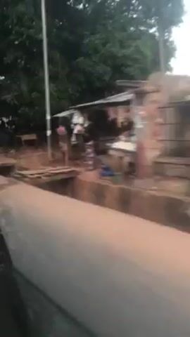 The video shows a scene from a moving vehicle, likely a car or bus, driving along a street. The street is lined with buildings, including small shops and houses, some of which appear to be in a state of disrepair. There are people visible along the roadside, some walking, some standing. The overall impression is of a modest, possibly low-income area. The road surface is visible, appearing to be paved. The weather seems overcast, contributing to the somewhat muted and dreary atmosphere of the scene.